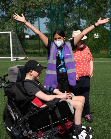 women in purple hi-vis jacket standing outside on grass with arms in the air next to a young boy with a black cap on in a black wheelchair