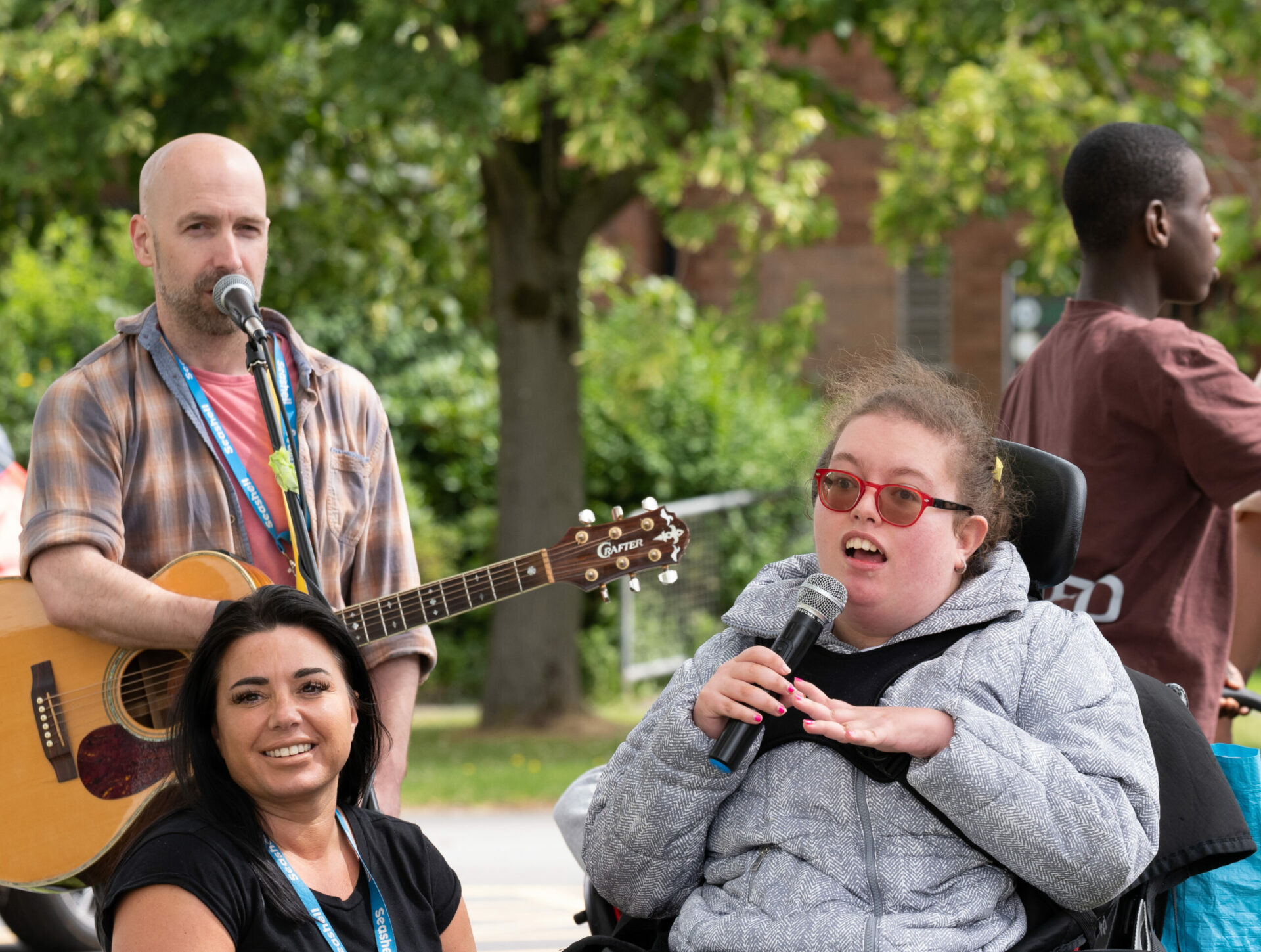 man in striped top stood holding guitar next to a girl sat in a wheelchair wearing a grey top and holding a microphone with seashell worker kneeling down next to her