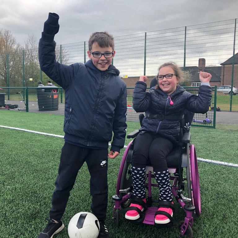 A boy and a girl cheering on a football pitch. The girl is in a wheelchair. 